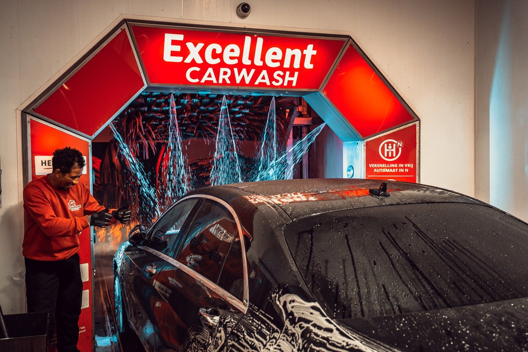 Medewerker van Excellent Carwash voert de handmatige voorwas uit onder de rode entree van de wasstraat aan de Langsom 2 in Amsterdam.